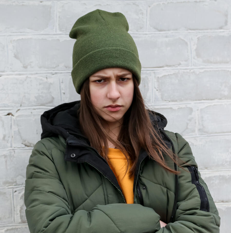 Teenage girl dressed in orange tp and green coat and hat. She is upset or grumpy but looking at the photographer. Her arms are folded. She may be suffering from anger, anxiety or depression.