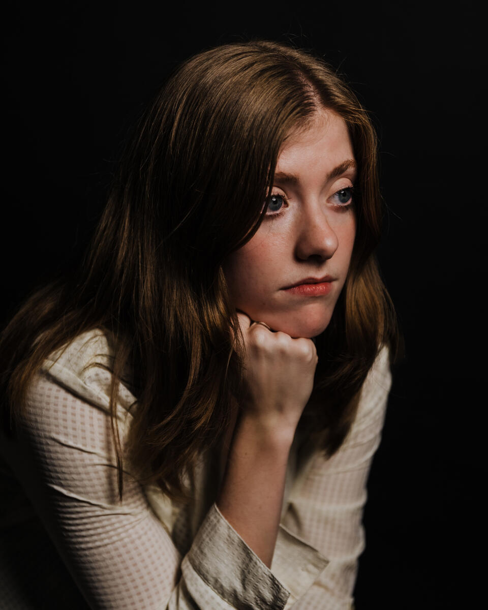 Photograph of a teenage girl with beautiful big eyes and lovely brown hair looking into the distance to the left of the camera with her chin resting on her hand. There is sadness in her face and she doesn't look interested in anyone or anything. She may be