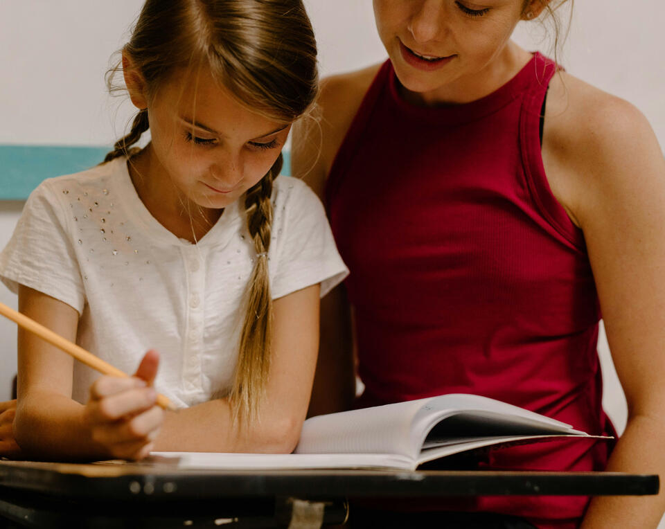 Photograph of mum supporting girl to do schoolwork at home. The girl is holding a pencil & ready to write or draw